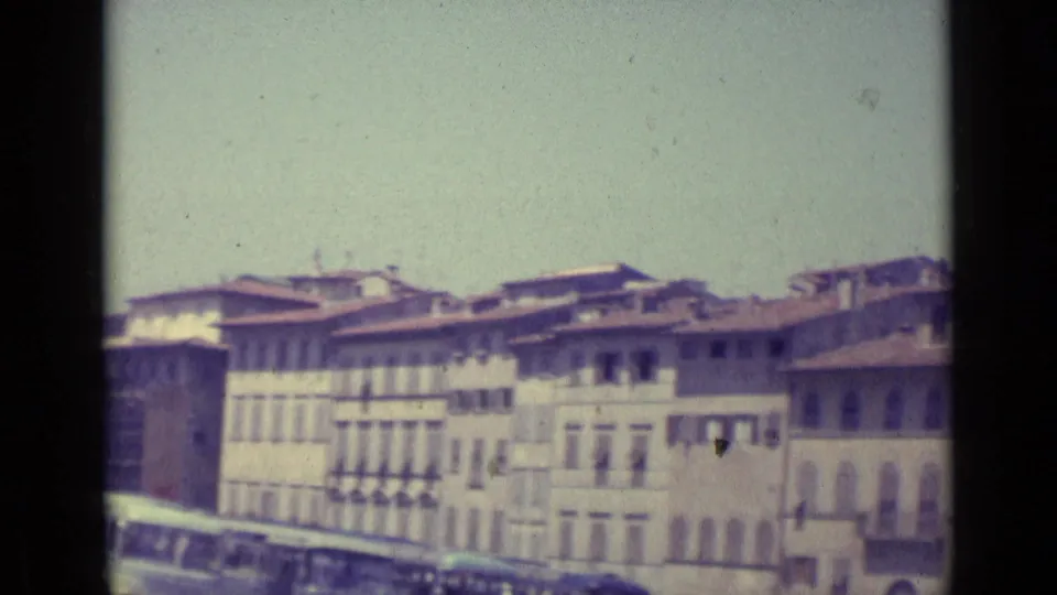 1962 Florence city view with pale building facades, terracotta roofs, shuttered windows, and a brief glimpse of vehicles at street level.
