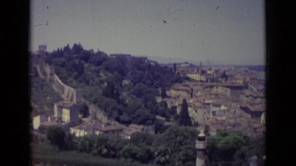 1962 Florence panorama from an elevated viewpoint, with green hillside, rooftops, river corridor, and distant urban skyline.