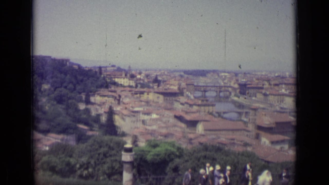 1962 Florence panorama with the Arno River, historic bridges, dense rooftops, trees, and tourists at the lower edge of frame.