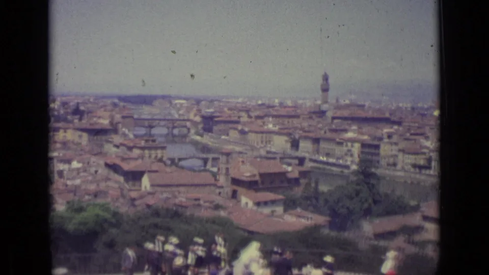 1962 Florence city panorama showing the Arno River, bridge crossings, red roofs, a tall tower, greenery, and visitors near the overlook.
