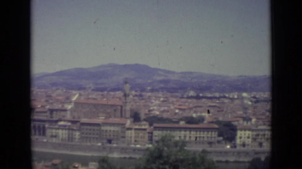 Elevated 1962 8mm view across Florence, with terracotta rooftops, riverfront buildings, a church tower, and hazy hills beyond.