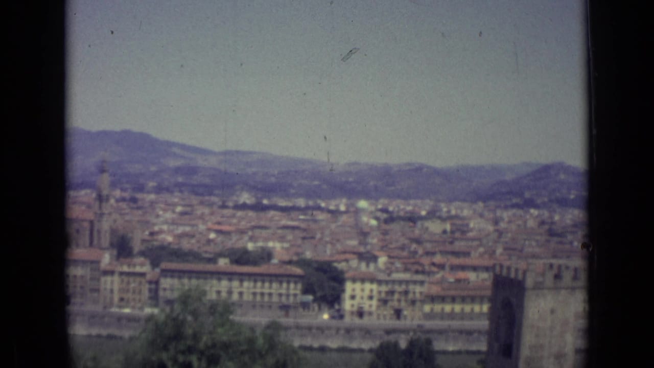 1962 home-movie frame looking across Florence, showing a broad cityscape, a prominent tower, the riverfront, and Tuscan hills under haze.