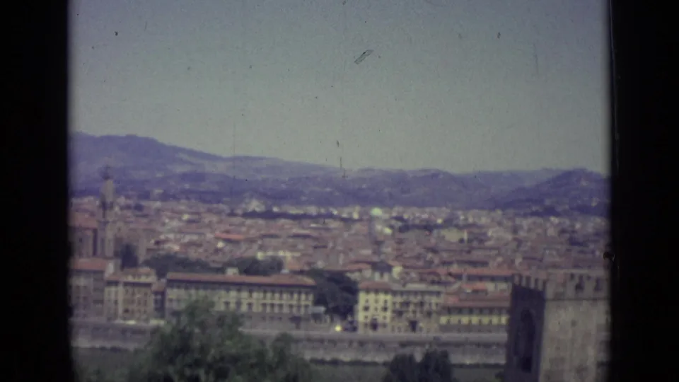 1962 home-movie frame looking across Florence, showing a broad cityscape, a prominent tower, the riverfront, and Tuscan hills under haze.