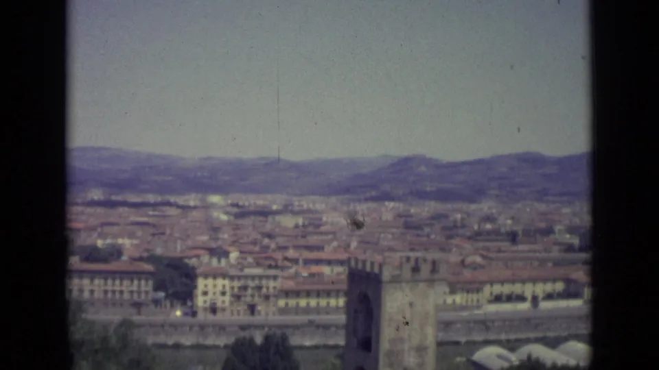 Mid-pan frame from a 1962 Florence overlook, with dense urban roofs, riverside architecture, hills, and a tower near the right edge.