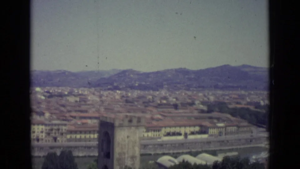 Later 1962 8mm frame of Florence from above, showing a tower, riverfront buildings, clustered roofs, low hills, and pale arched roofs below.