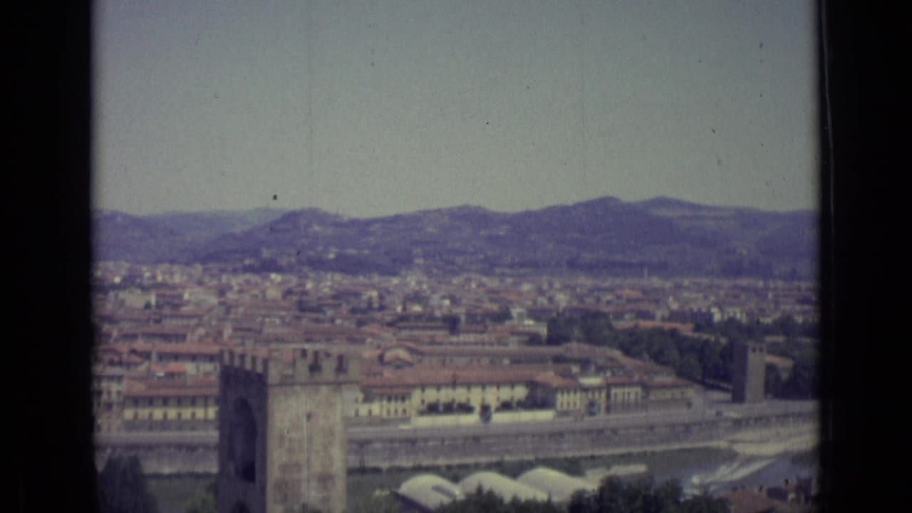 Elevated view over Florence with a stone tower, river, city buildings, hills, and pale sky.