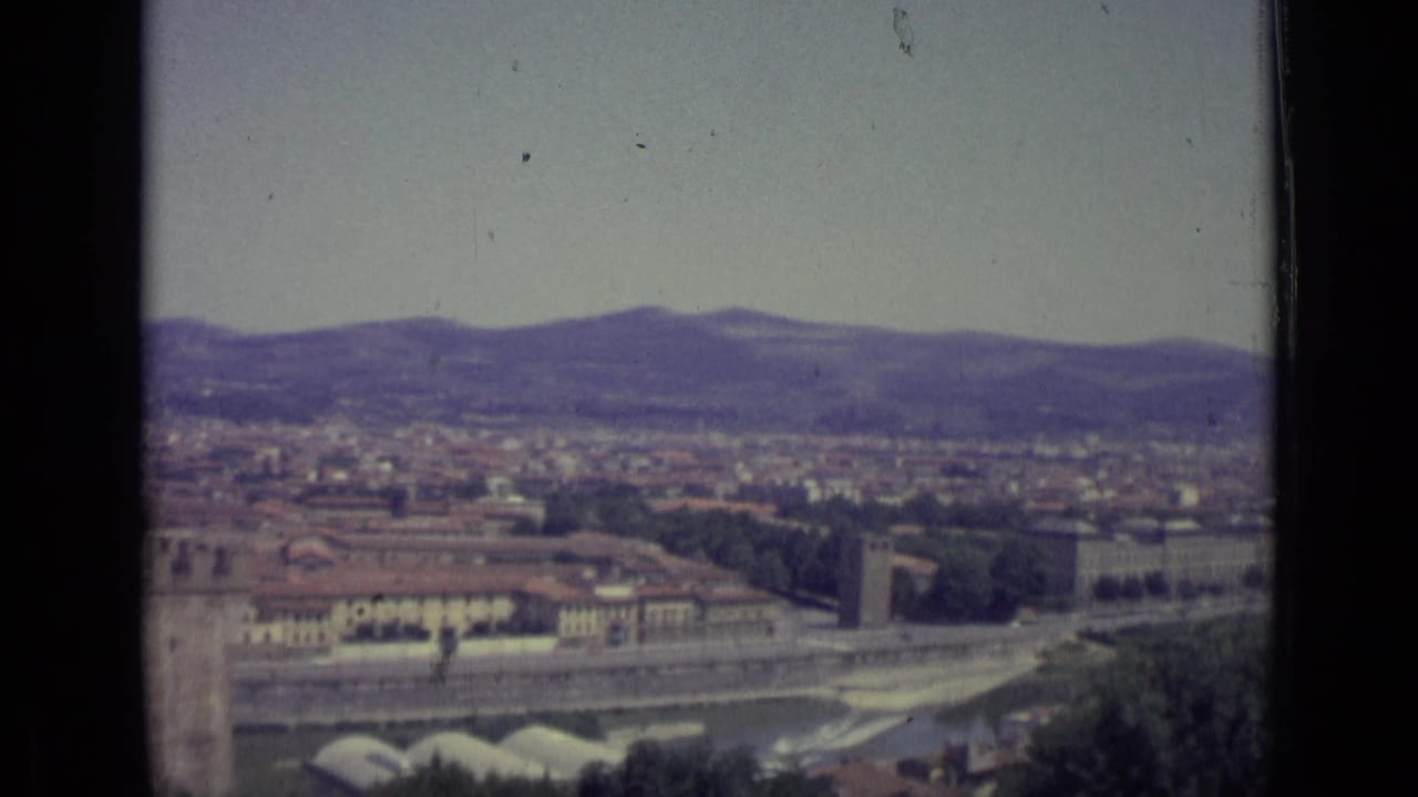 Florence cityscape seen from above with river embankment, rooftops, trees, hills, and sky.