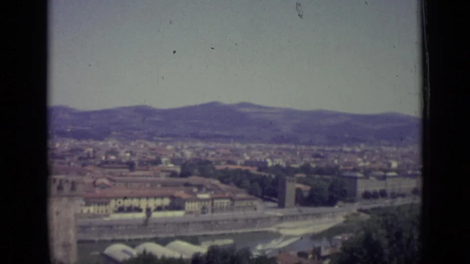 1962 home-movie frame of Florence from an elevated overlook, showing the river corridor, clustered rooftops, trees, distant hills, and faded sky.