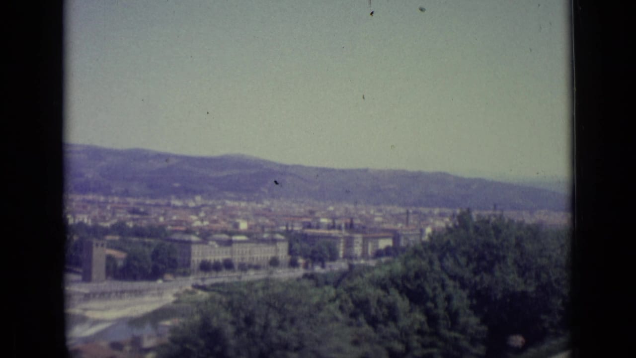 Wide view over a city with a river, trees in the foreground, and hills under a hazy sky.