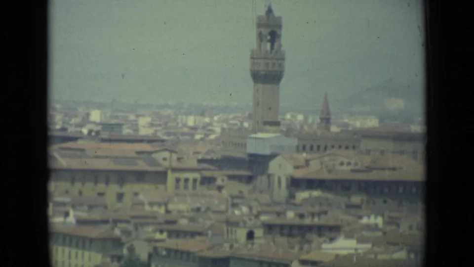 1962 8mm skyline view of Florence with the Palazzo Vecchio tower, historic rooftops, and a distant church spire in haze.