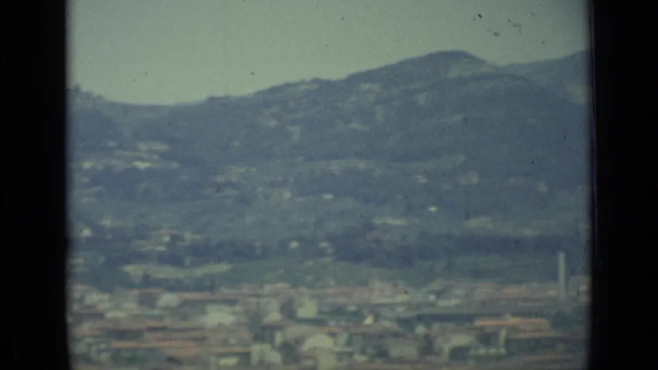1962 Florence home-movie frame showing a soft, distant cityscape beneath tree-covered hills and a pale sky.
