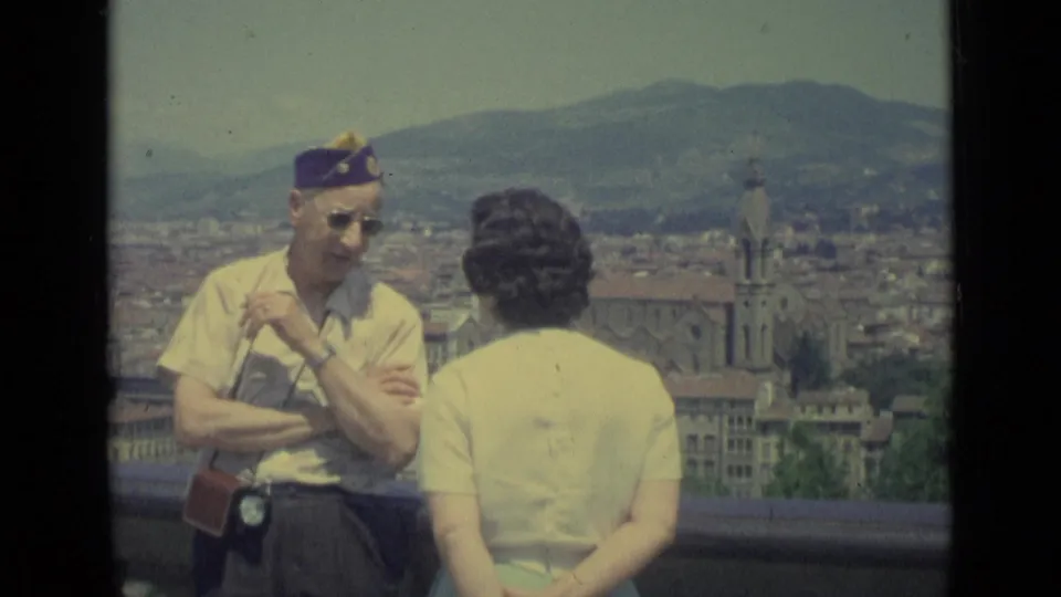 In 1962 Florence, an adult man in sunglasses and a purple cap talks with a woman at an overlook above the city.