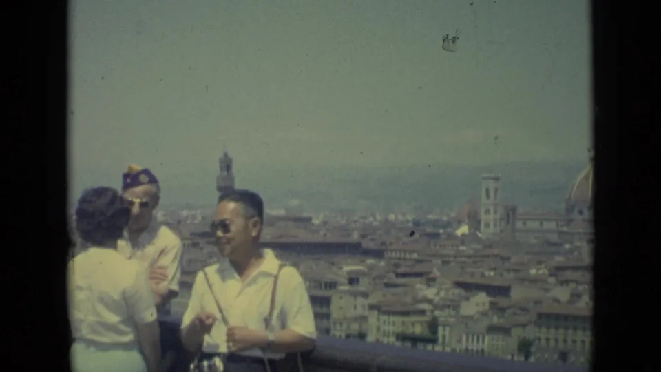 1962 Florence home-movie frame showing adults talking and posing at a viewpoint, with the Duomo dome and campanile visible across the city.