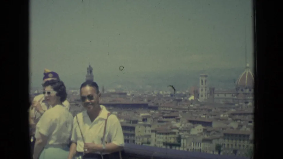 1962 Florence home-movie frame showing smiling adult tourists at an overlook, with the city skyline, campanile, and edge of the Duomo behind them.