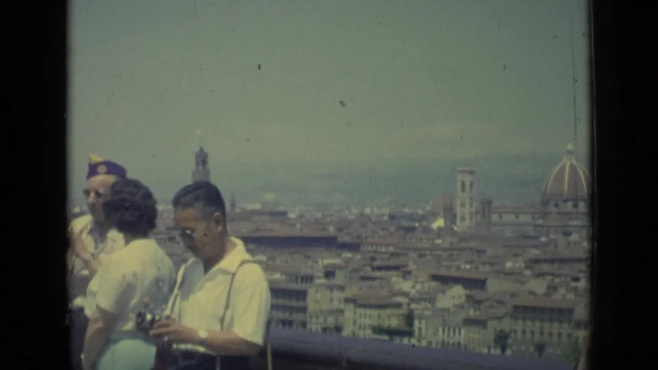 1962 Florence terrace view with tourists, a handheld camera, rooftops, bell towers, and the Duomo dome in the background.