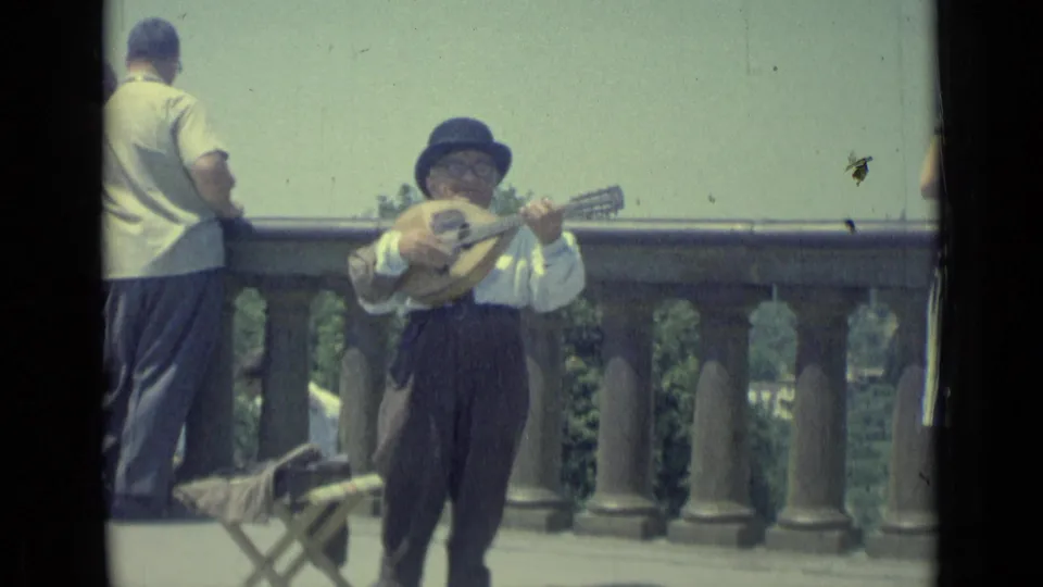 1962 Florence home-movie frame of a street musician performing with a mandolin-like instrument near a stone terrace railing.