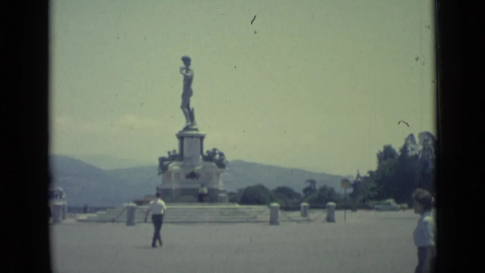 1962 Florence home-movie frame showing a public monument in a plaza, with a pedestrian near the base and another person at frame edge.