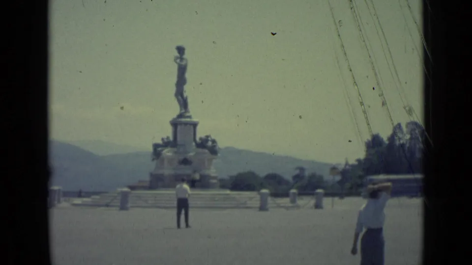 Florence 1962 8mm frame with the monument centered, a man near the plaza middle, and a woman passing close to the camera.