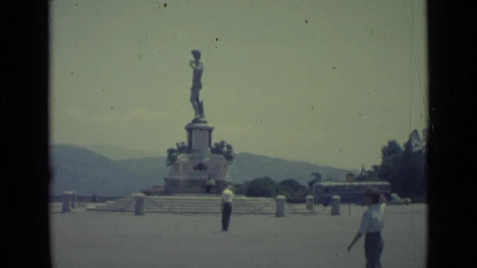 1962 Florence home-movie frame showing the monument, a pedestrian in the plaza, and a bus moving behind the statue.