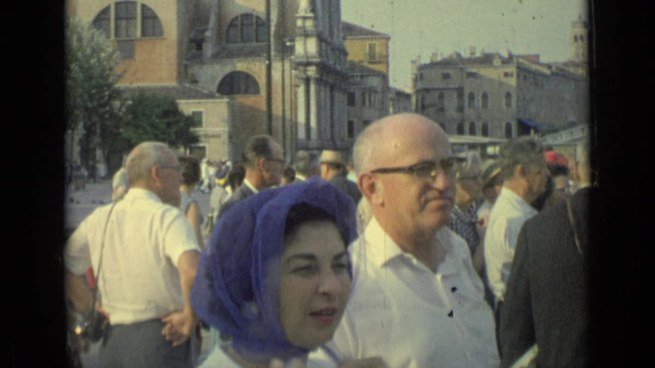 Crowd of adults near historic arched buildings, with a woman in a blue headscarf in the foreground.