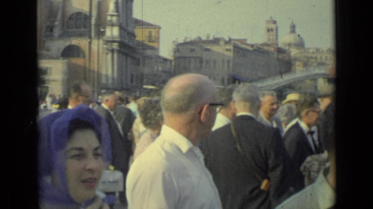 Adults stand in a dense crowd near arched buildings and a bridge-like structure in the background.
