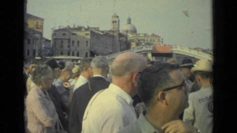 1962 8mm frame of a crowded Italian public location, with men in glasses, uniformed attendants and historic buildings behind a bridge.