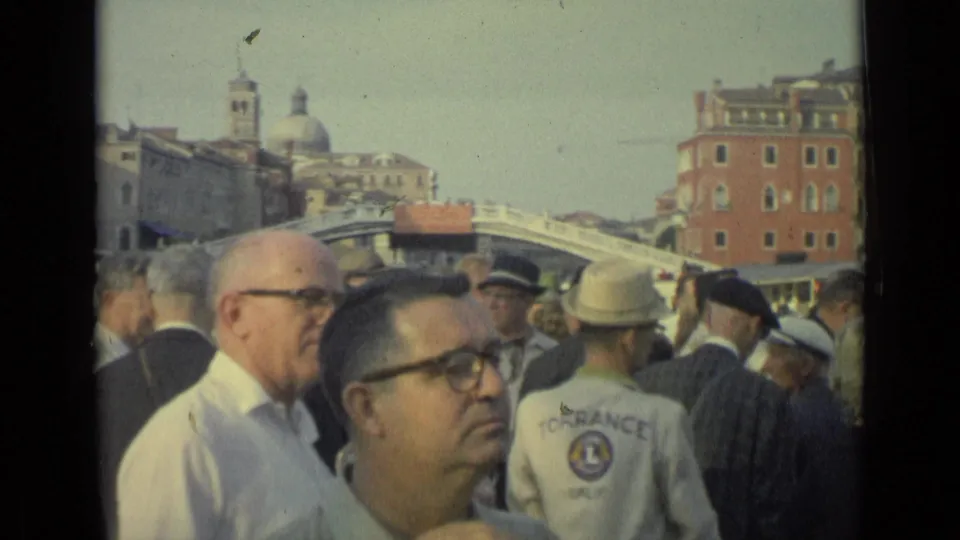 1962 Italy crowd footage showing a foreground man with glasses, a bald man in a white shirt, uniformed attendants and a bridge beyond.