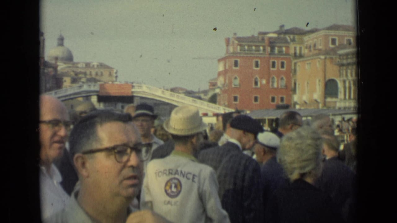 Crowd of tourists standing near water with arched bridge and Italian buildings behind them.
