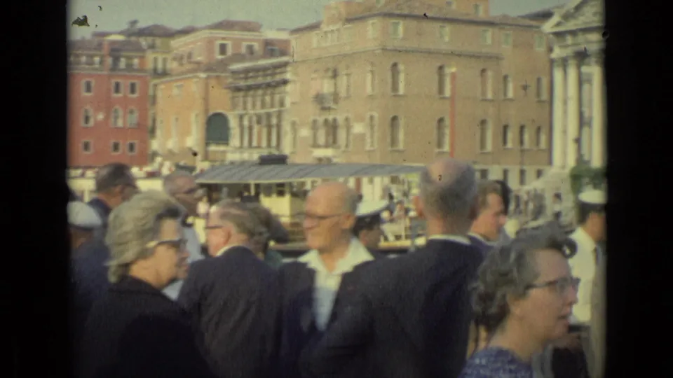 1962 Florence home-movie frame of tourists near the water, with boats, awnings, and pale building facades beyond them.