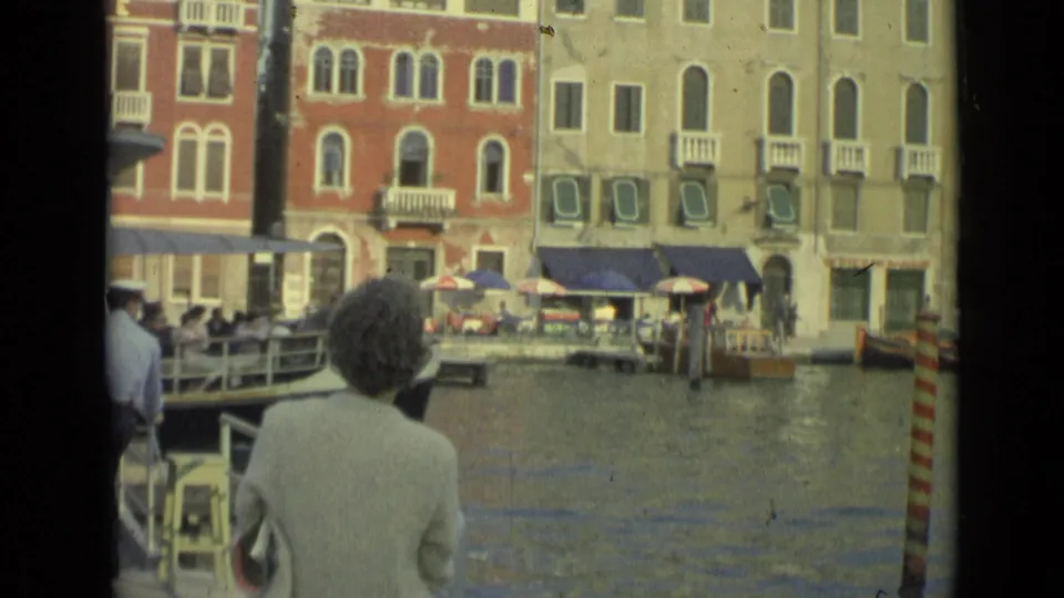 1962 Florence waterfront frame with a woman facing the water, striped mooring poles, cafe umbrellas, and multi-story facades.
