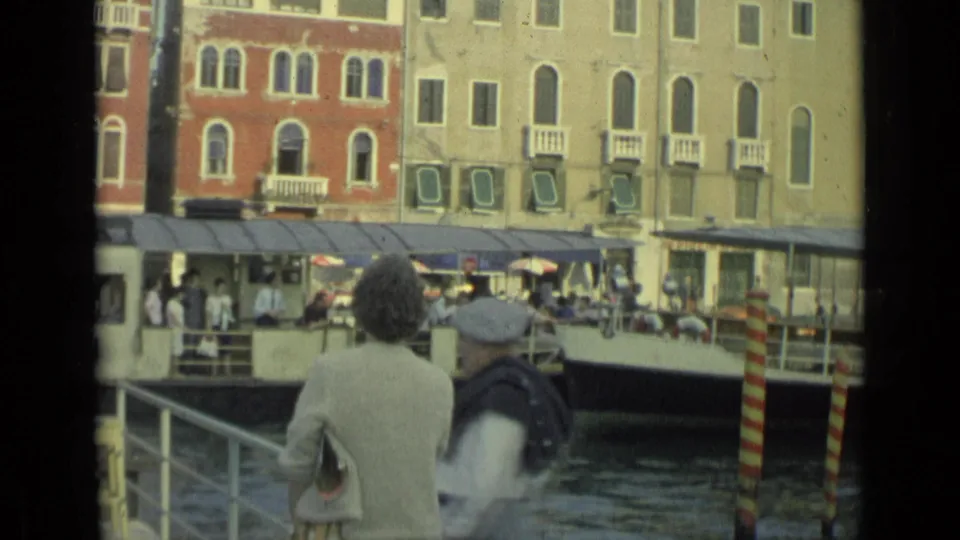 A larger canal passenger boat crosses the frame near a woman at the dock, with cafe awnings, striped poles, and old Italian buildings behind it.