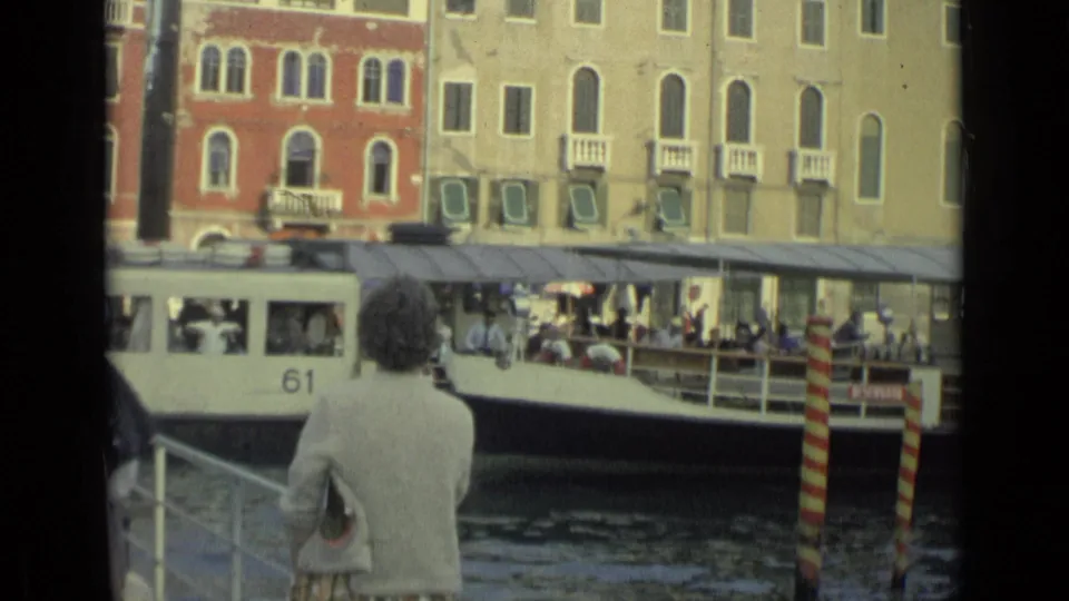 A white passenger boat with the number 61 passes a woman on a canal dock, with striped mooring poles and cafe seating in the background.