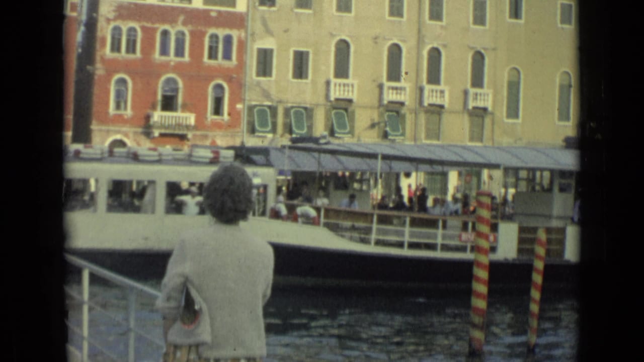 Woman stands by a canal railing as a white passenger boat passes historic waterfront buildings.