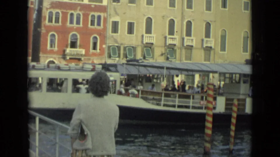 In archival 1962 Italian home-movie footage, a woman faces a canal while a white public boat passes in front of arched buildings.