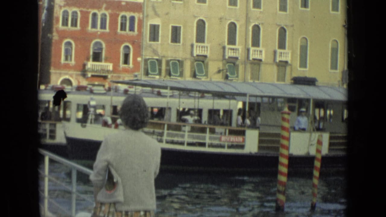 The woman remains at the canal edge while a vaporetto-style passenger boat and dock structure pass across the 1962 home-movie frame.