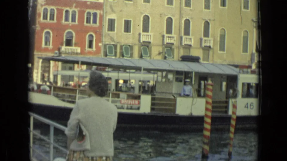 A white canal passenger boat with visible passengers passes close to the dock as the woman observes in 1962 Italian home-movie footage.
