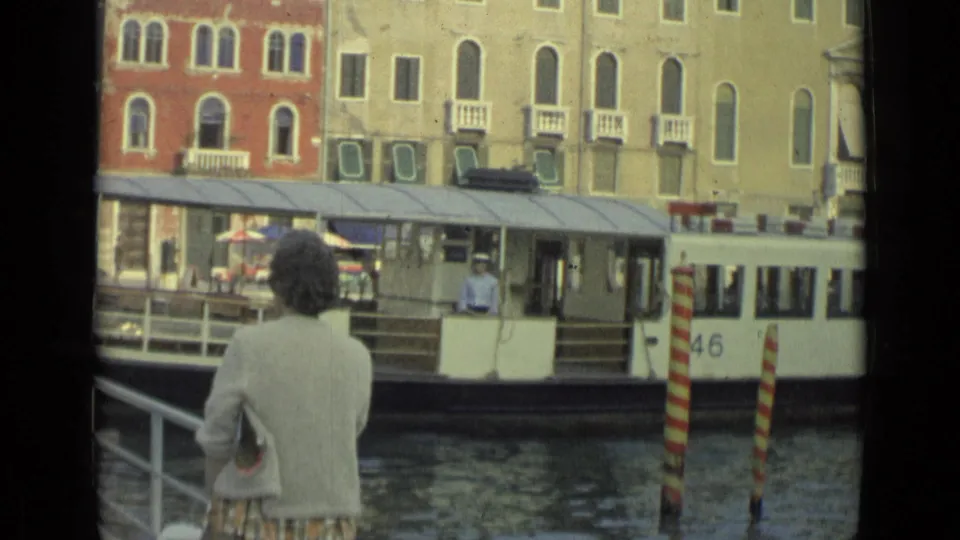 The frame shows a woman watching a white public canal boat pass between striped posts and waterfront buildings in archival 1962 footage.