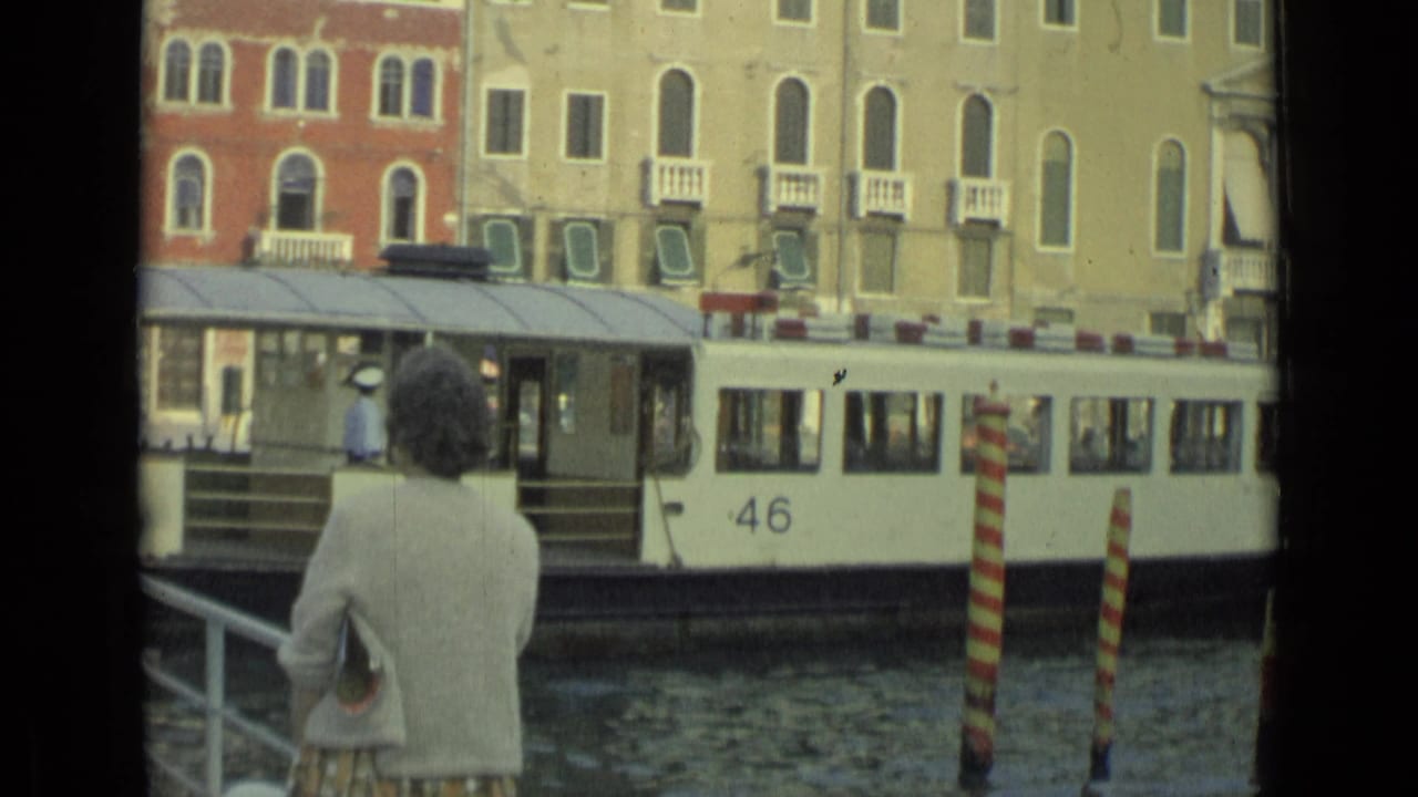 A woman in a light sweater watches a white water bus marked 46 pass along an Italian canal in 1962 footage.