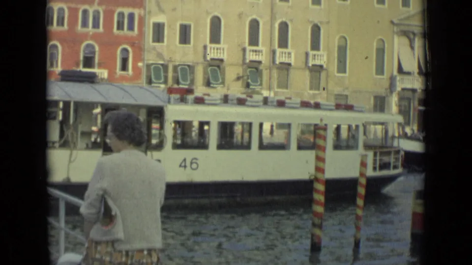 The home-movie frame shows a dockside woman facing a passing white canal water bus, with Venetian-style facades behind it.