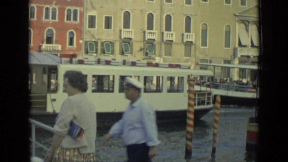 A uniformed man passes in front of a woman at the canal railing while the white water bus continues behind them.
