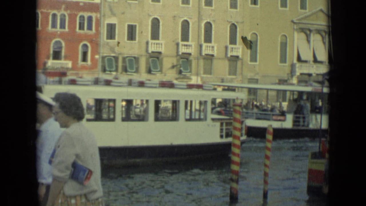 Woman near a canal as a white passenger boat passes striped mooring poles.