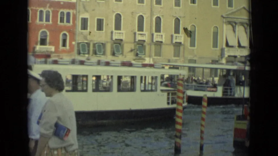 1962 Italian 8mm travel frame showing a pedestrian beside canal water while a white public boat passes shuttered buildings.