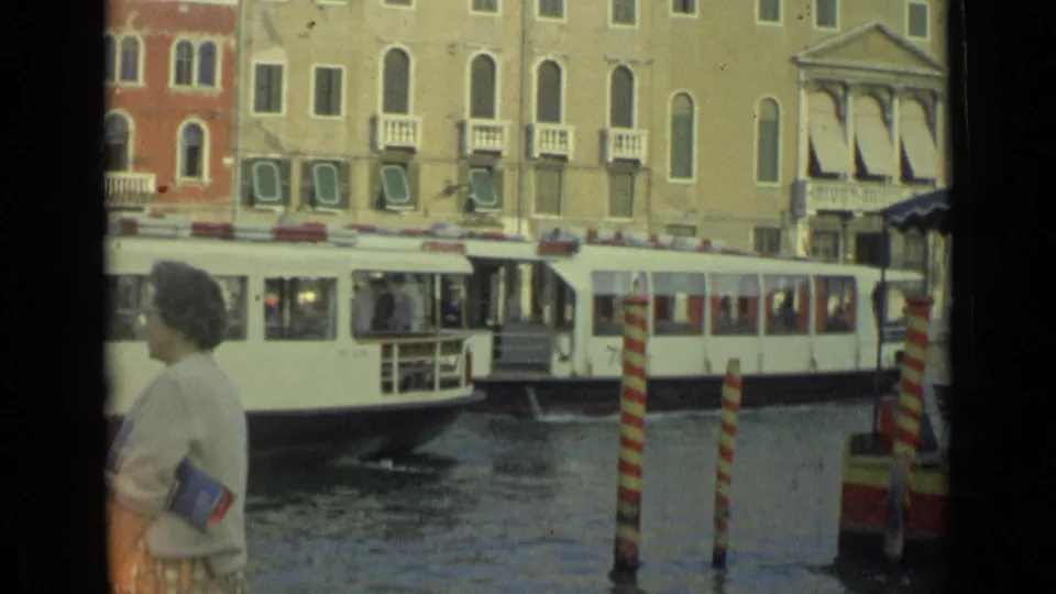 1962 8mm frame of a canal-side pedestrian with a white passenger boat moving across the background in Italy.