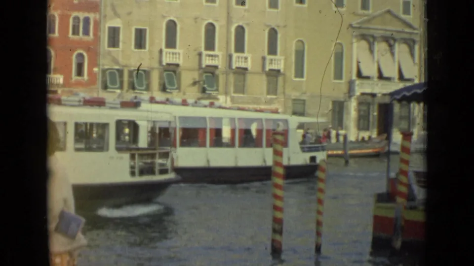 1962 Italian canal view with a white passenger boat, striped mooring poles, waterfront facades, and a smaller boat at right.