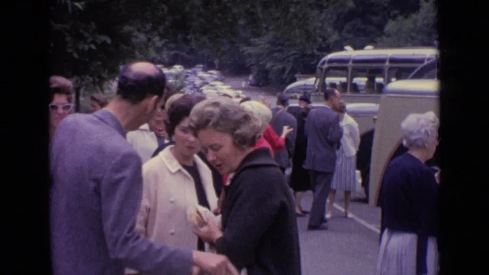 Adults in mid-century coats and suits gather near vintage buses on a wooded roadside in Germany, 1962.