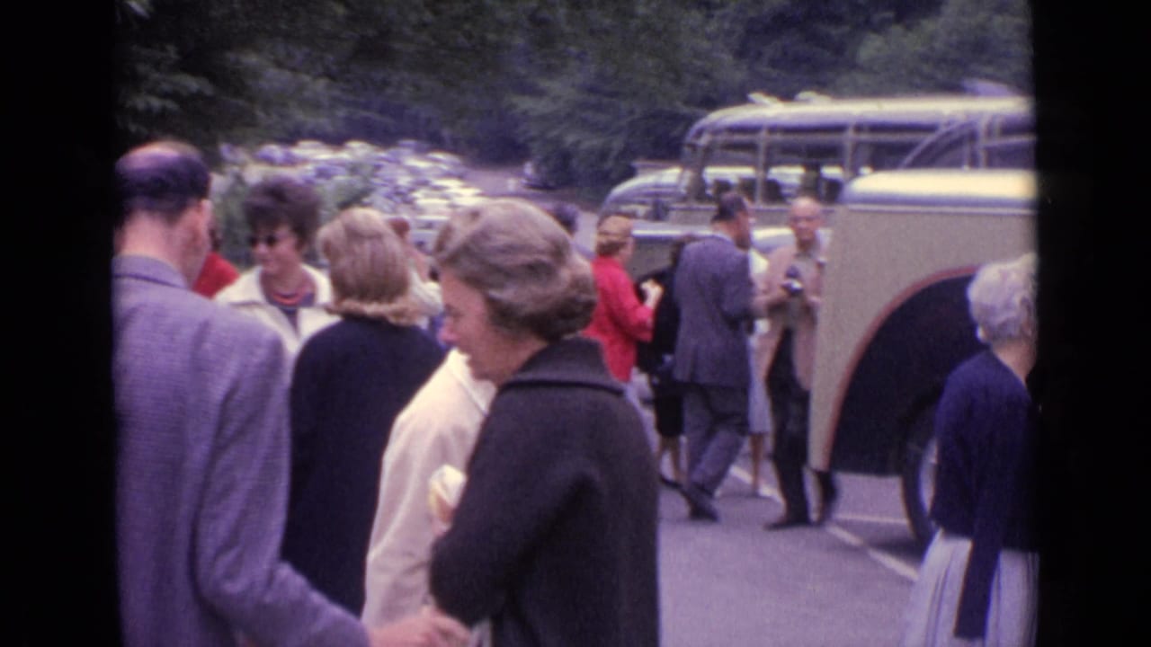 1962 Germany home-movie frame showing adults mingling in a parking lot near cream-and-dark coach buses.