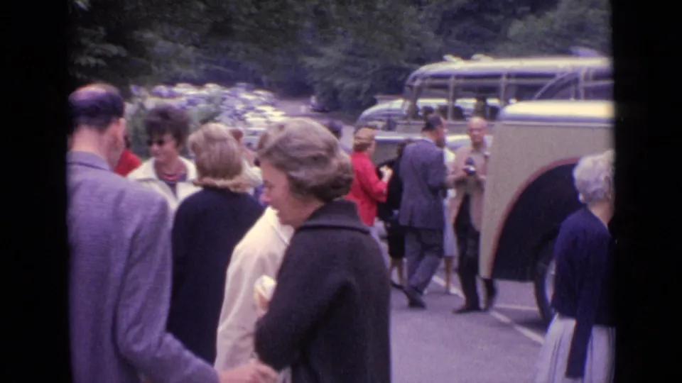 1962 Germany home-movie frame showing adults mingling in a parking lot near cream-and-dark coach buses.