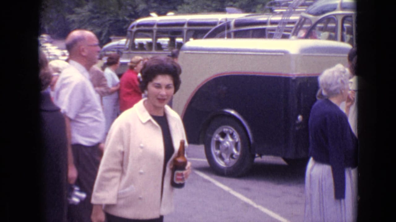 A woman in a cream jacket walks through a parking lot holding a beer bottle near a coach bus.