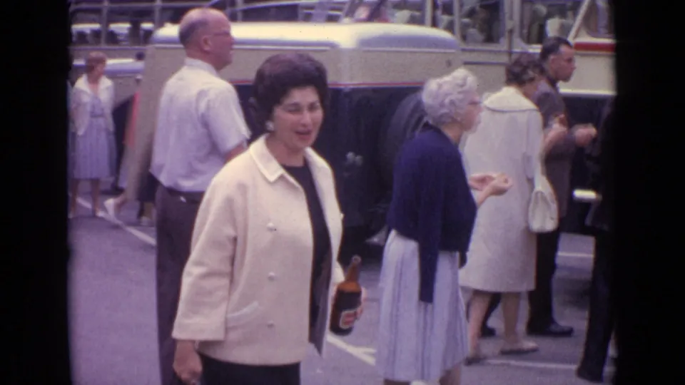 Home-movie view of the cream-jacketed woman approaching the camera while people gather near a vintage bus in Germany.
