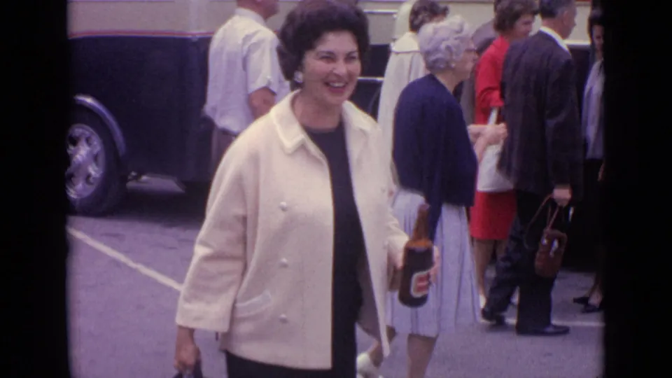 Smiling woman carrying a beer bottle in a busy 1962 Germany parking-lot scene with adults and a coach bus behind her.
